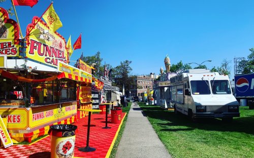 Colorful food stalls line a sunny pathway, featuring funnel cakes and other treats, with blue skies above.