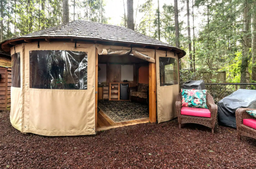 A cozy yurt surrounded by trees, featuring large windows and a small porch with outdoor seating.
