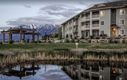 A modern building by a pond, reflecting mountains and clouds in the background, surrounded by greenery.
