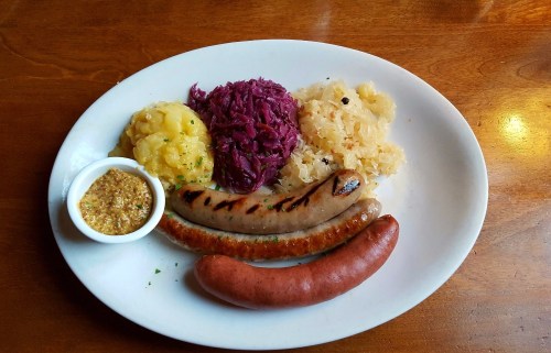 A plate with two types of sausages, mashed potatoes, red cabbage, sauerkraut, and a small bowl of mustard.