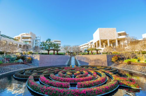 A landscaped garden with a floral maze, water features, and modern buildings under a clear blue sky.