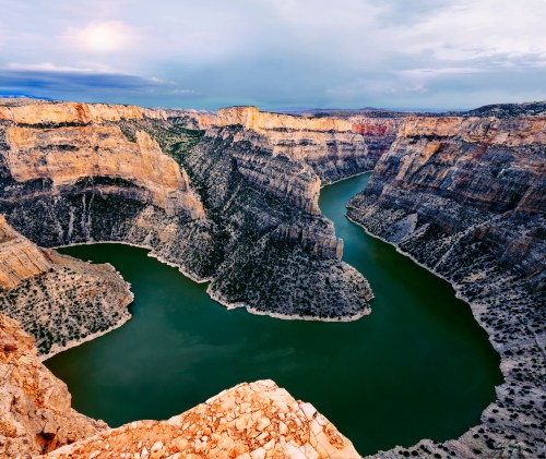 Aerial view of a winding river surrounded by steep, rocky cliffs and lush greenery under a cloudy sky.