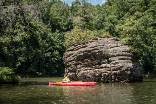 A person in a red kayak paddles near a large rock formation surrounded by lush green trees and calm water.