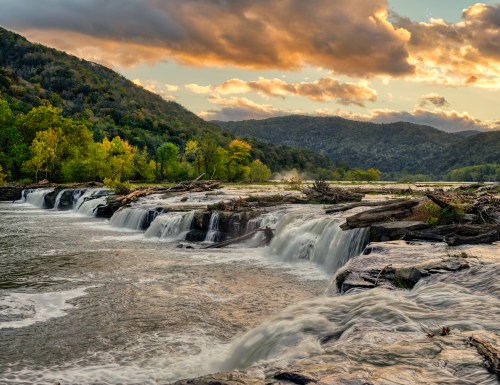 A serene waterfall cascades over rocks, surrounded by lush greenery and mountains under a colorful sunset sky.