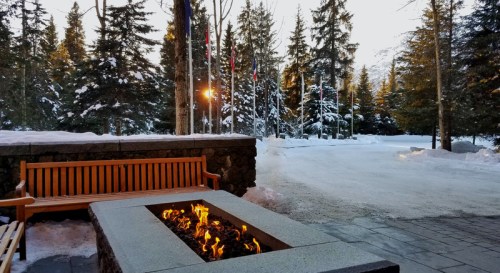 A cozy outdoor seating area with a fire pit, surrounded by snow-covered trees and flags, at sunset.