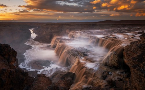 A stunning waterfall cascades over rocky cliffs at sunset, surrounded by mist and dramatic clouds in the sky.