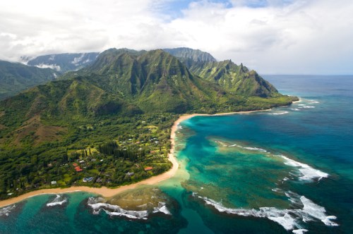 Aerial view of a lush green coastline with mountains, clear blue waters, and sandy beaches under a cloudy sky.