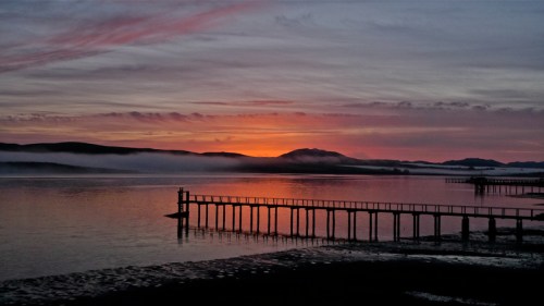 A serene sunset over calm waters, with a silhouetted pier and misty hills in the background.