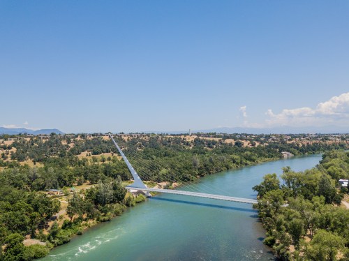 Aerial view of a suspension bridge over a river, surrounded by green trees and rolling hills under a clear blue sky.