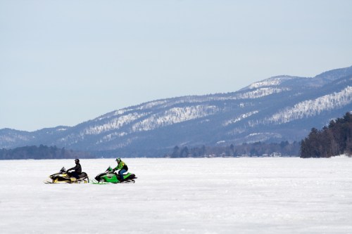 Two snowmobiles ride across a snowy landscape with mountains in the background on a clear winter day.
