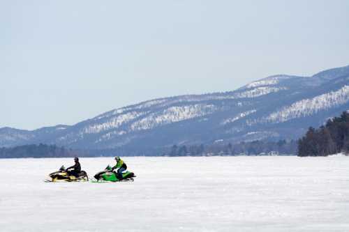 Two snowmobiles ride across a snowy landscape with mountains in the background on a clear winter day.