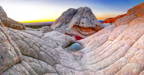 A panoramic view of unique white rock formations at sunset, with a small pool of water reflecting the colors of the sky.