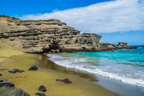 A scenic beach with golden sand, rocky cliffs, and turquoise waves under a partly cloudy sky.