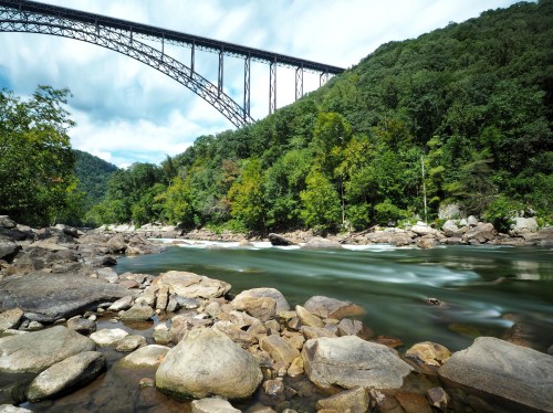 A flowing river with smooth stones, framed by lush greenery and a towering bridge above against a cloudy sky.