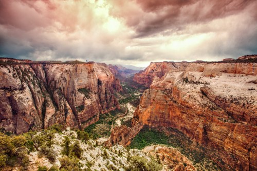 A dramatic view of Zion Canyon with towering cliffs, a winding river, and a moody sky filled with clouds.