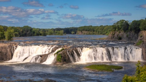 A scenic view of a waterfall cascading over rocks, surrounded by lush greenery and a blue sky with scattered clouds.