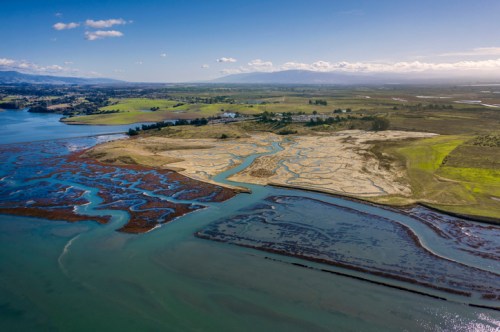 Aerial view of a coastal landscape with winding waterways, marshlands, and green fields under a clear blue sky.