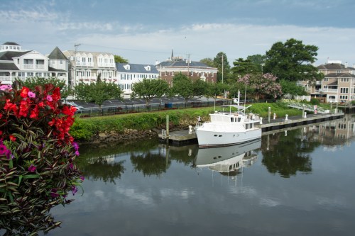 A serene waterfront scene with a white boat docked beside colorful flowers and charming houses reflected in the water.