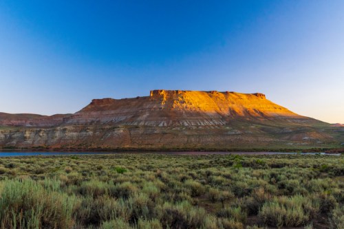 A sunlit plateau rises above a grassy plain, with a clear blue sky in the background and a river at the base.