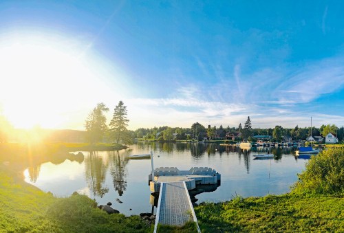 A serene lakeside view at sunset, featuring a dock, boats, and trees reflecting on calm water.