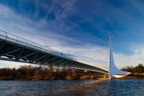 A modern cable-stayed bridge spans a river, with a striking white tower and blue sky in the background.