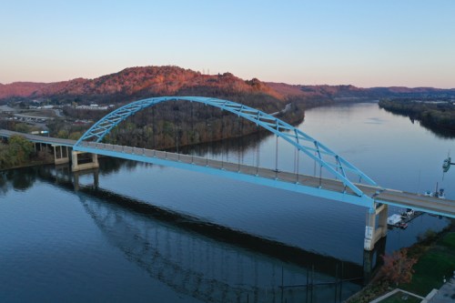 A blue arch bridge spans a calm river, reflecting in the water, with hills in the background at sunset.