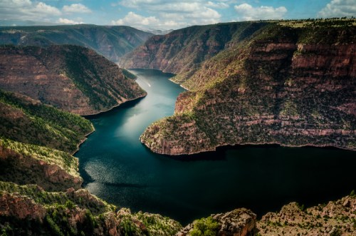 A scenic view of a winding river surrounded by steep, rocky cliffs and lush greenery under a partly cloudy sky.