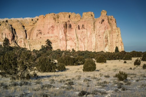 A rugged cliff with pink and beige rock formations towers over a grassy landscape dotted with evergreen trees.