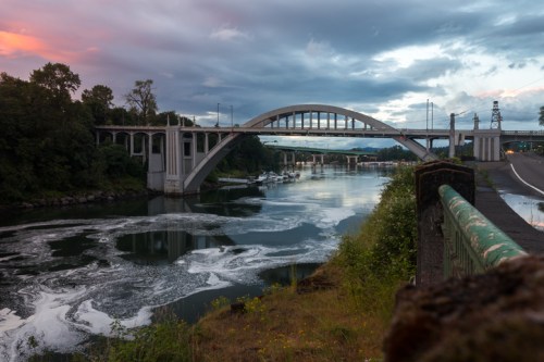A bridge arches over a river, reflecting clouds and trees, with a road alongside and foamy water below.
