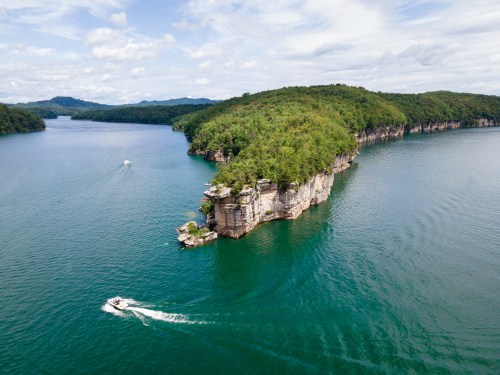 Aerial view of a serene lake with a rocky island and boats navigating the water, surrounded by lush greenery.
