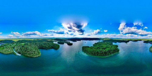 A panoramic view of a serene lake surrounded by lush green islands and a bright blue sky with scattered clouds.