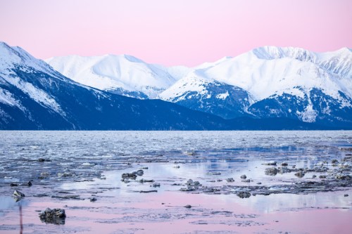 Snow-capped mountains reflect in a frozen lake at sunset, with a soft pink sky and ice fragments scattered on the water.