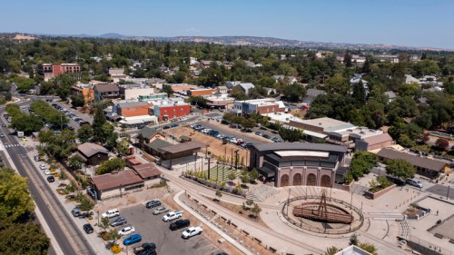 Aerial view of a small town with shops, trees, and a central plaza featuring a circular structure and parking areas.