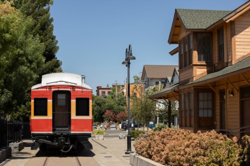 A red train car parked beside a wooden station, surrounded by trees and colorful buildings under a clear blue sky.