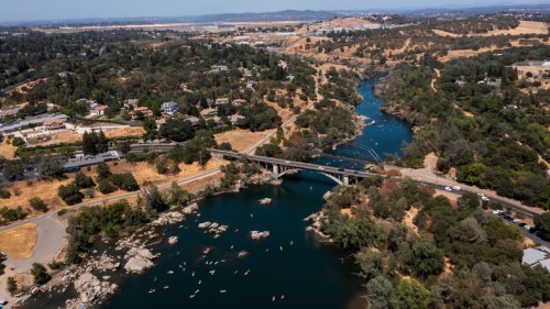 Aerial view of a river winding through green hills, with a bridge and kayakers visible along the water's edge.