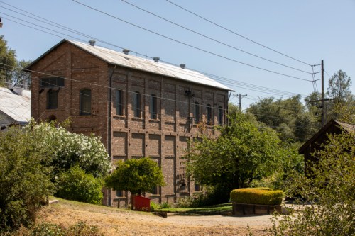 A brick building surrounded by greenery, with power lines in the background under a clear blue sky.