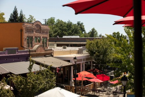 A view of a vibrant outdoor dining area with red umbrellas, surrounded by trees and historic buildings.