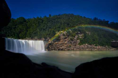 A serene waterfall cascades into a calm river, with a vibrant rainbow arching over lush green cliffs under a starry sky.