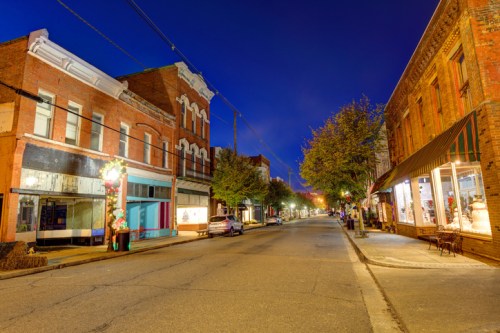 Quiet street scene at dusk, featuring brick buildings, trees, and storefronts illuminated by soft streetlights.