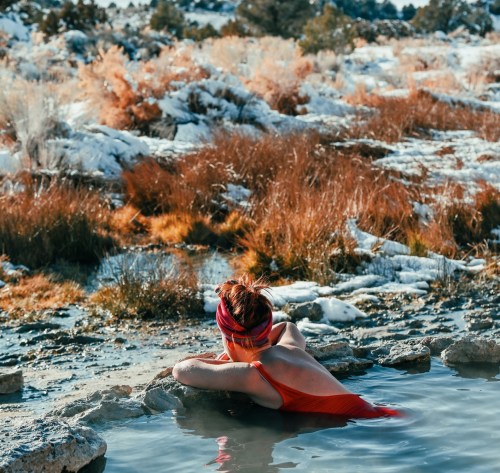 A person in a red swimsuit relaxes in a natural hot spring surrounded by snowy landscapes and autumn foliage.