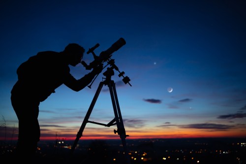 A silhouette of a person using a telescope against a twilight sky with stars and a crescent moon.