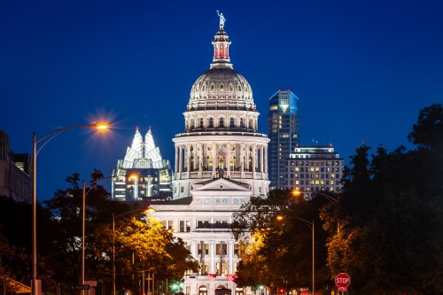 The Texas State Capitol illuminated at night, with modern buildings in the background and streetlights in the foreground.