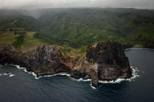 Aerial view of a rugged coastline with cliffs and lush green hills under a cloudy sky. Ocean waves crash against the shore.