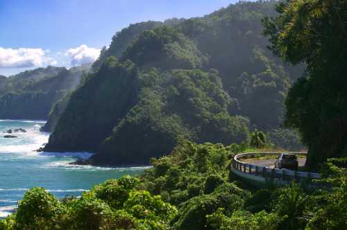 A scenic coastal road winds along lush green hills, with waves crashing against the shore below.