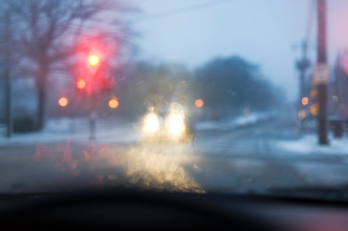 Blurry view from a car during a snowy night, with red traffic light and headlights illuminating the road ahead.