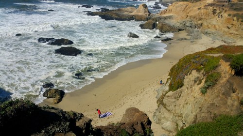 A scenic beach with waves crashing on rocks, two people on the sand, and cliffs covered in greenery in the background.