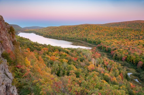 A scenic view of a river winding through a vibrant autumn forest, with colorful foliage and rolling hills under a pastel sky.