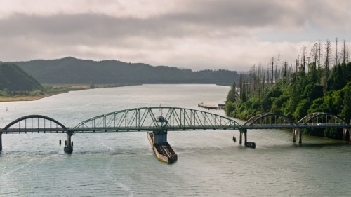 A green bridge spans a river, surrounded by lush greenery and distant hills under a cloudy sky.