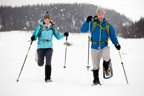 Two people joyfully snowshoeing through a snowy landscape, wearing winter gear and smiling. Snowflakes are falling around them.