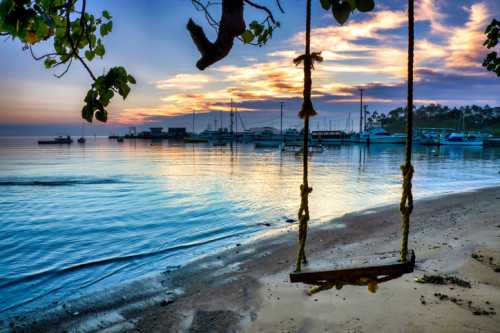 A serene beach at sunset with a swing hanging from a tree, boats docked in the background, and calm water reflecting the sky.